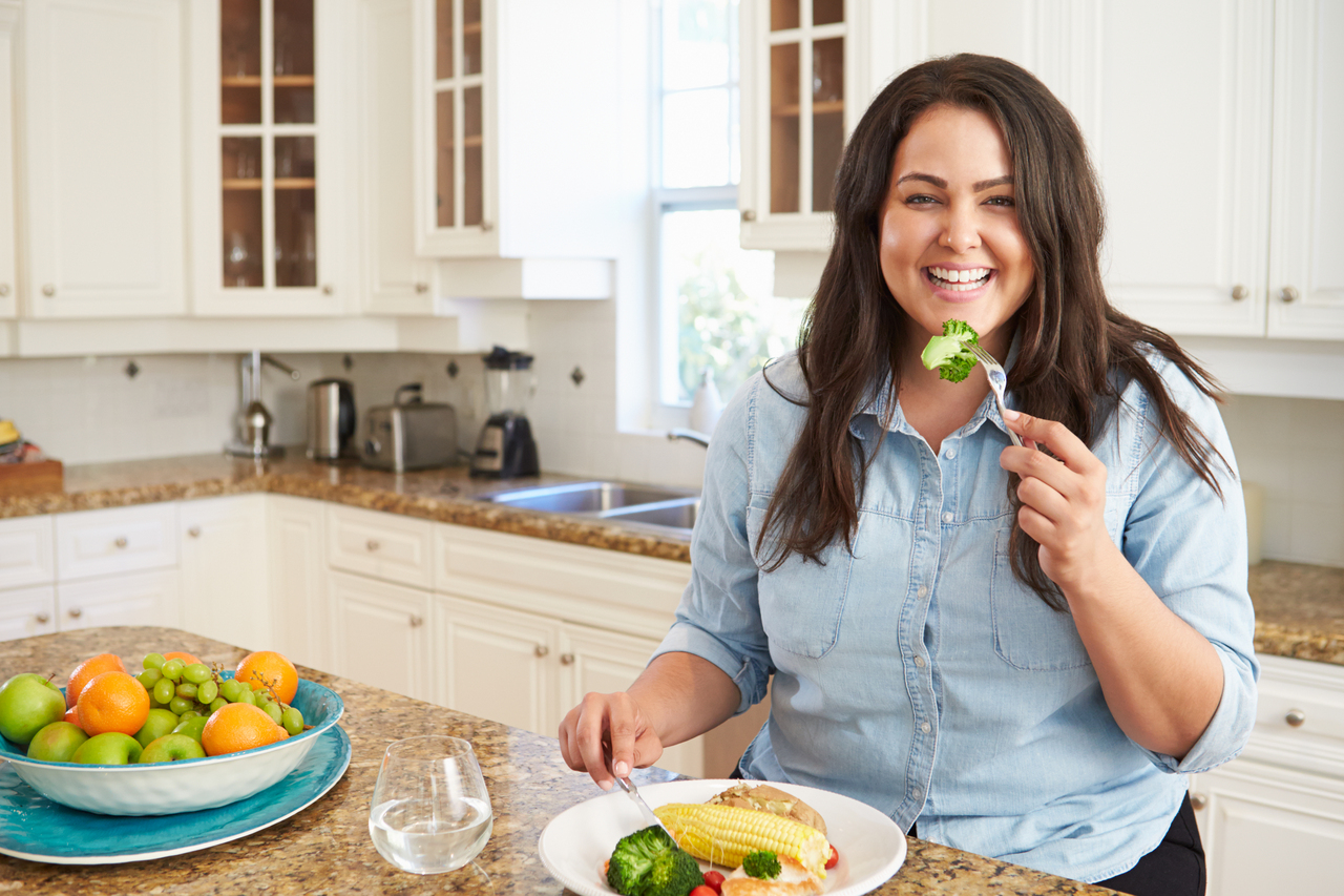 Overweight Woman Eating Healthy Meal In Kitchen Sitting Down Smiling At Camera